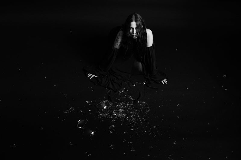 B/W image of Chelsea Wolfe in dark clothing crouching down near broken glass